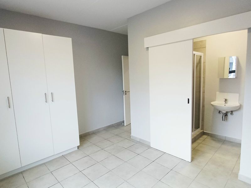 A clean, empty bedroom featuring built-in cupboards, tiled flooring, and a sliding door leading to an en-suite bathroom with a shower and basin.