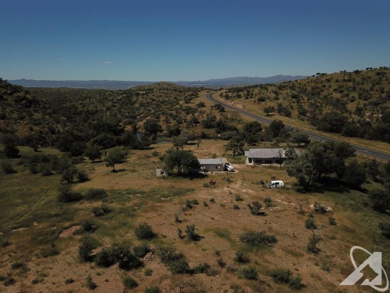 Aerial view of a large vacant residential plot featuring natural landscape, existing structures, and proximity to a main road.