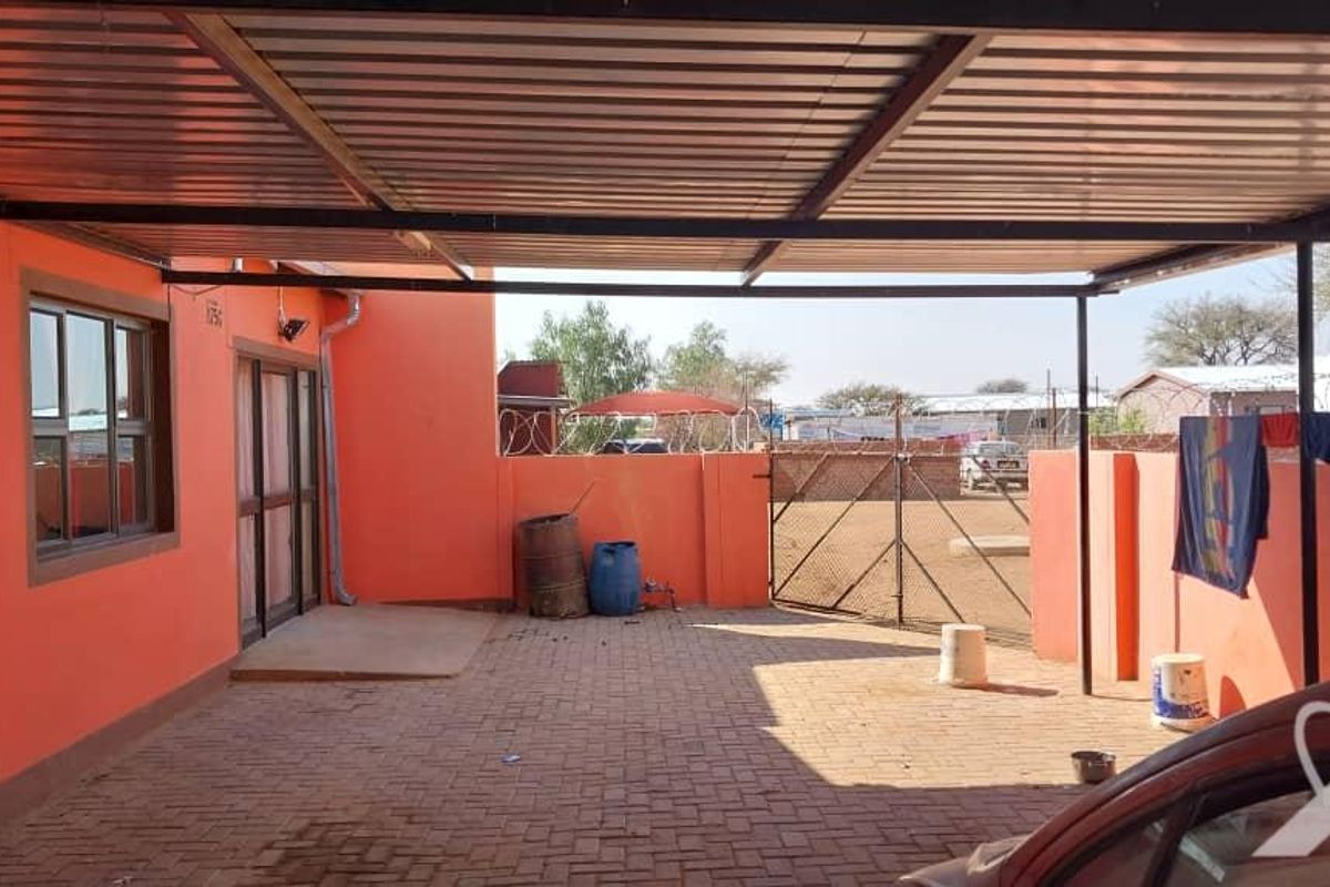 A covered outdoor patio area featuring paved flooring, a metal roof structure, and perimeter boundary walls with security fencing.