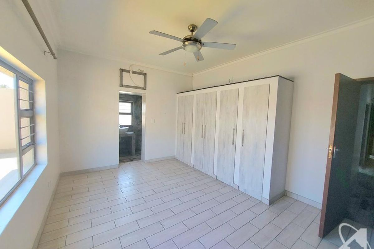 A bright, empty bedroom featuring tiled flooring, built-in cupboards, a ceiling fan, and an en-suite bathroom entrance.