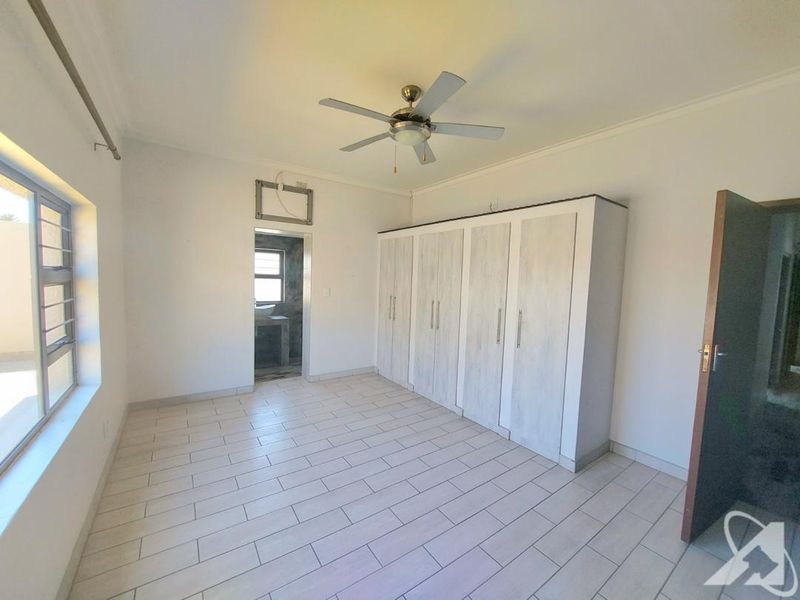 A bright, empty bedroom featuring tiled flooring, built-in cupboards, a ceiling fan, and an en-suite bathroom entrance.