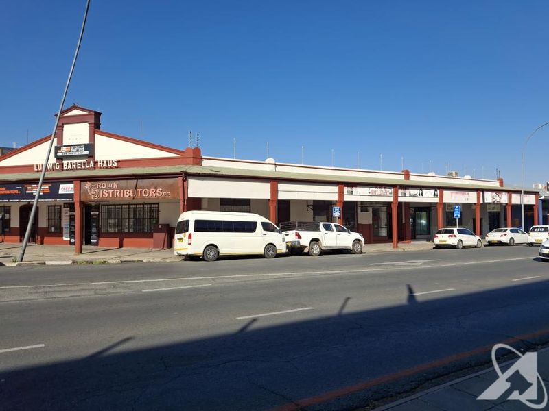 Street-level view of a commercial building facade featuring multiple storefronts, signage, and security fencing along a public road.