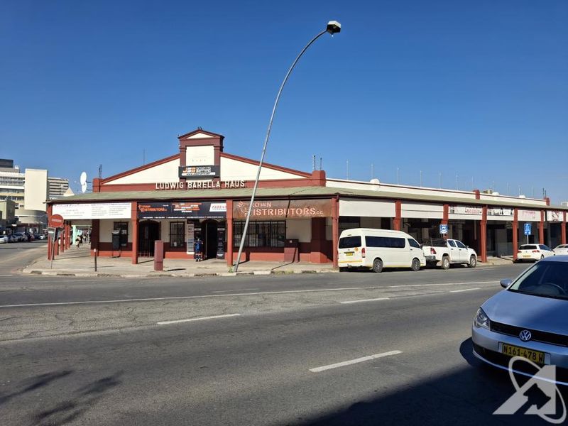 Street-level view of Ludwig Barella Haus, a commercial building featuring a covered walkway, prominent signage, and secure perimeter fencing.