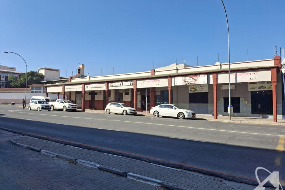 A street-level view of a commercial building featuring multiple storefronts with a covered walkway and paved sidewalk access.
