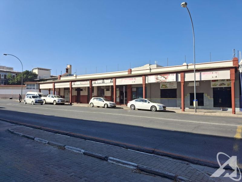 A street-level view of a commercial building featuring multiple storefronts with a covered walkway and paved sidewalk access.