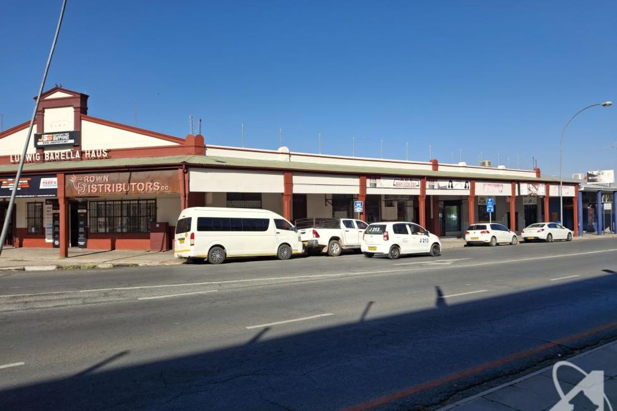 Street-level view of a commercial building featuring multiple storefronts, a covered walkway, and security fencing along the roofline.