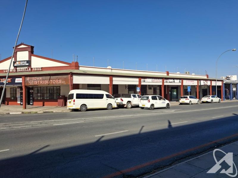 Street-level view of a commercial building featuring multiple storefronts, a covered walkway, and security fencing along the roofline.