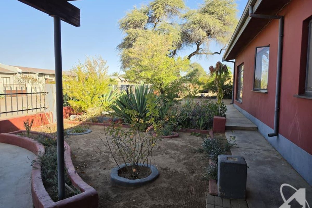 A rear exterior view featuring a covered patio area, concrete pathways, and established garden beds with raised borders alongside the house.