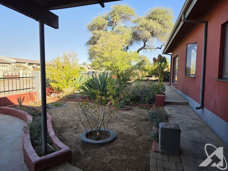 A rear exterior view featuring a covered patio area, concrete pathways, and established garden beds with raised borders alongside the house.