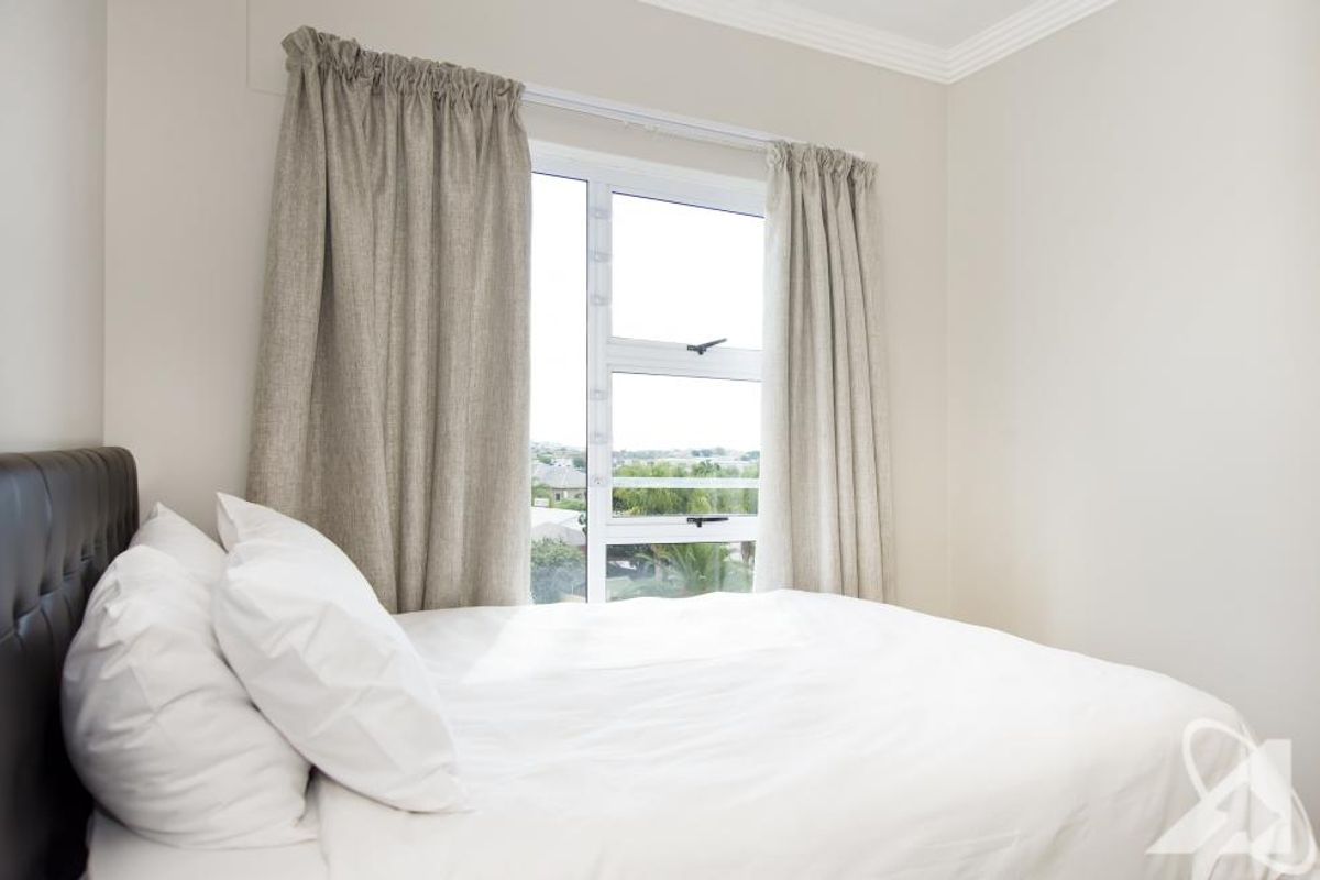 A bright bedroom featuring neutral walls, crown molding, and an aluminium window frame with curtains, offering a clean and simple living space.
