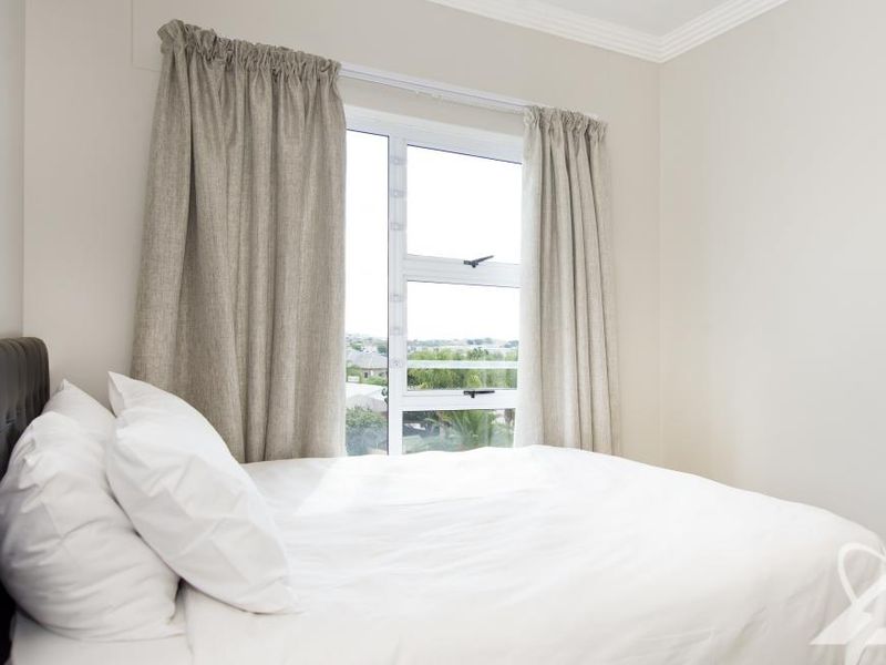 A bright bedroom featuring neutral walls, crown molding, and an aluminium window frame with curtains, offering a clean and simple living space.