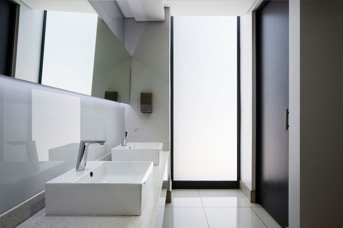 Modern commercial bathroom featuring dual vessel sinks, stone vanity countertop, large wall-mounted mirror, and frosted glass window for natural light.