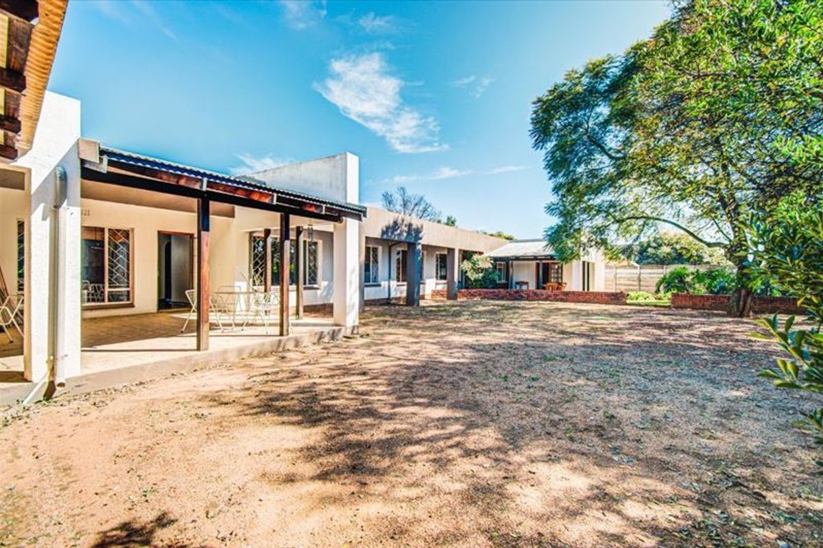 A spacious rear exterior view featuring a covered patio area with timber supports, overlooking a large garden space with mature trees.