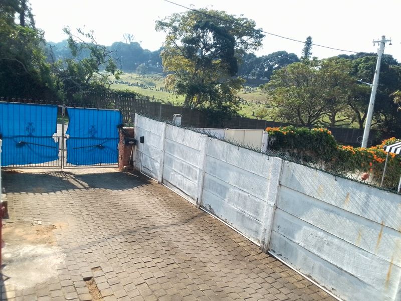 Front entrance area featuring a paved driveway, precast concrete boundary wall with security spikes, and a blue metal gate.
