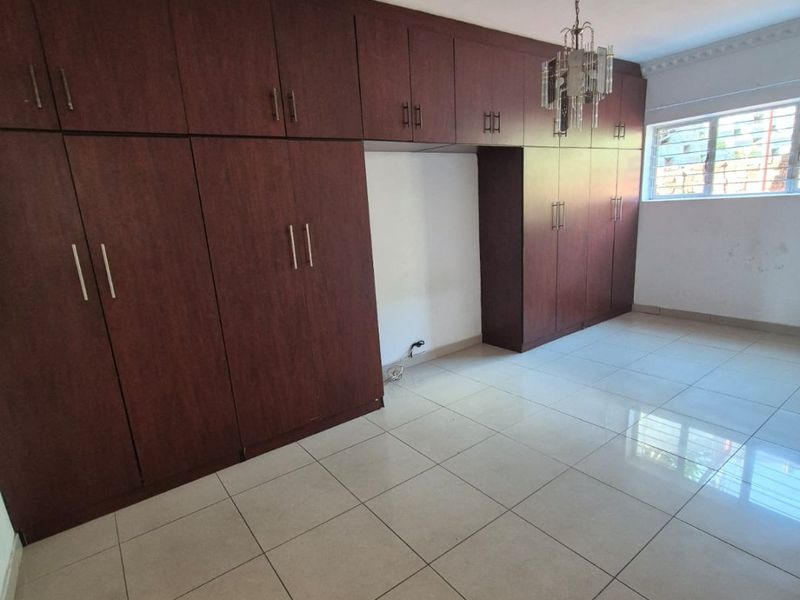 A spacious bedroom featuring extensive built-in wooden cupboards, neutral tiled flooring, and a window with security bars.
