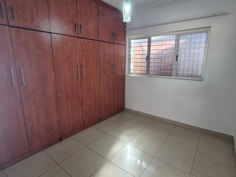 An empty bedroom featuring extensive built-in wooden cupboards, tiled flooring, and a window fitted with security bars.