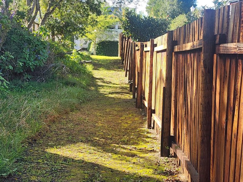 A narrow garden pathway bordered by a timber fence, showing signs of wear and uneven terrain.