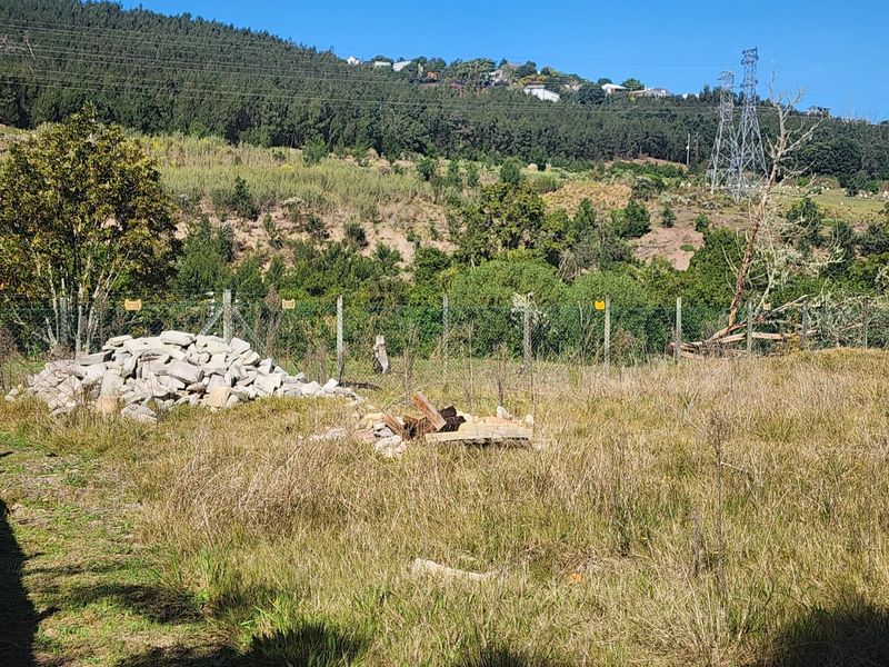 A view of a vacant residential plot featuring overgrown grass, a wire mesh fence, and surrounding hillside landscape.