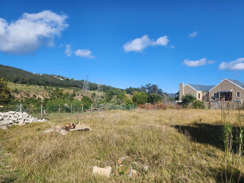 A vacant residential plot featuring overgrown grass, wire fencing, and scenic mountain views, offering potential for development in a quiet setting.
