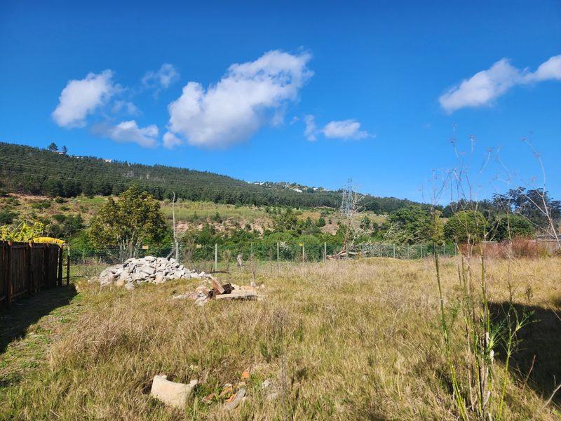 A vacant residential plot featuring overgrown grass, a wire mesh perimeter fence, and a wooden boundary fence with scenic mountain views.