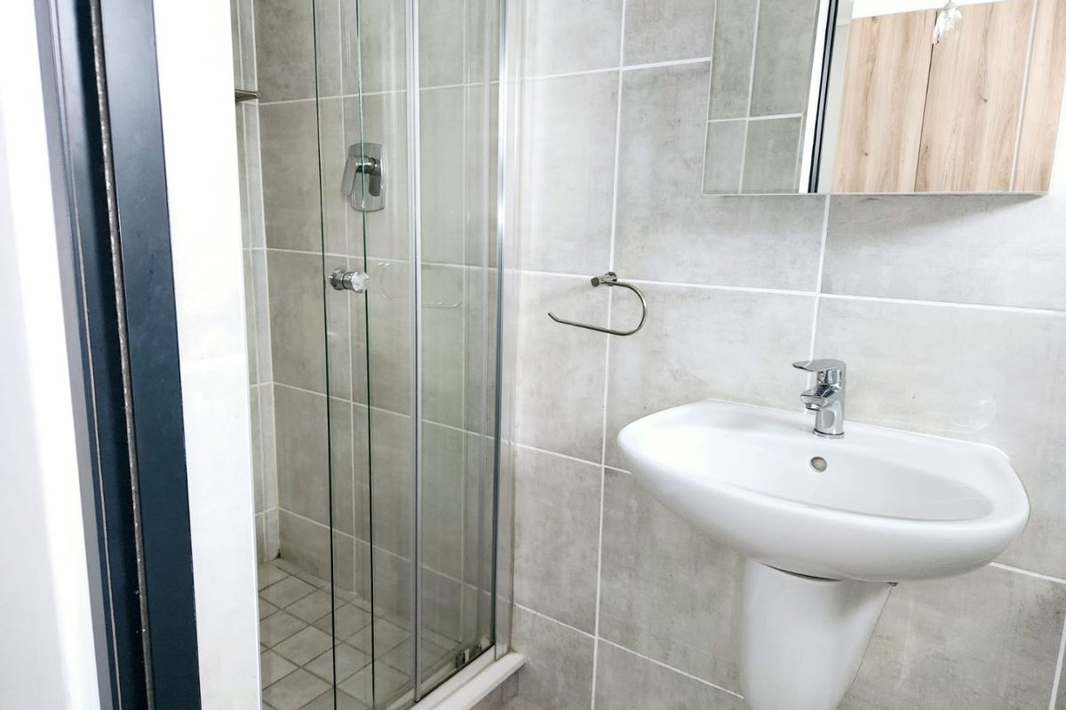 A modern, clean bathroom featuring a pedestal washbasin, chrome fixtures, and a glass-enclosed shower with neutral grey wall and floor tiling.