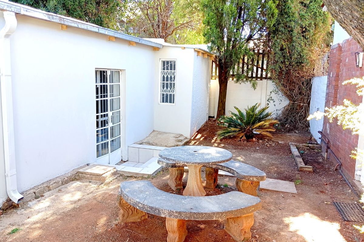 Rear exterior area featuring a white building facade, security-fitted doors and windows, and a garden space with a brick boundary wall.