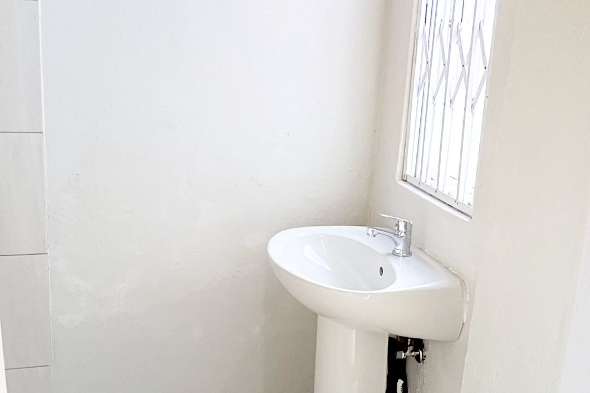 A compact bathroom featuring a white pedestal basin, tiled flooring, and a window fitted with a security gate.