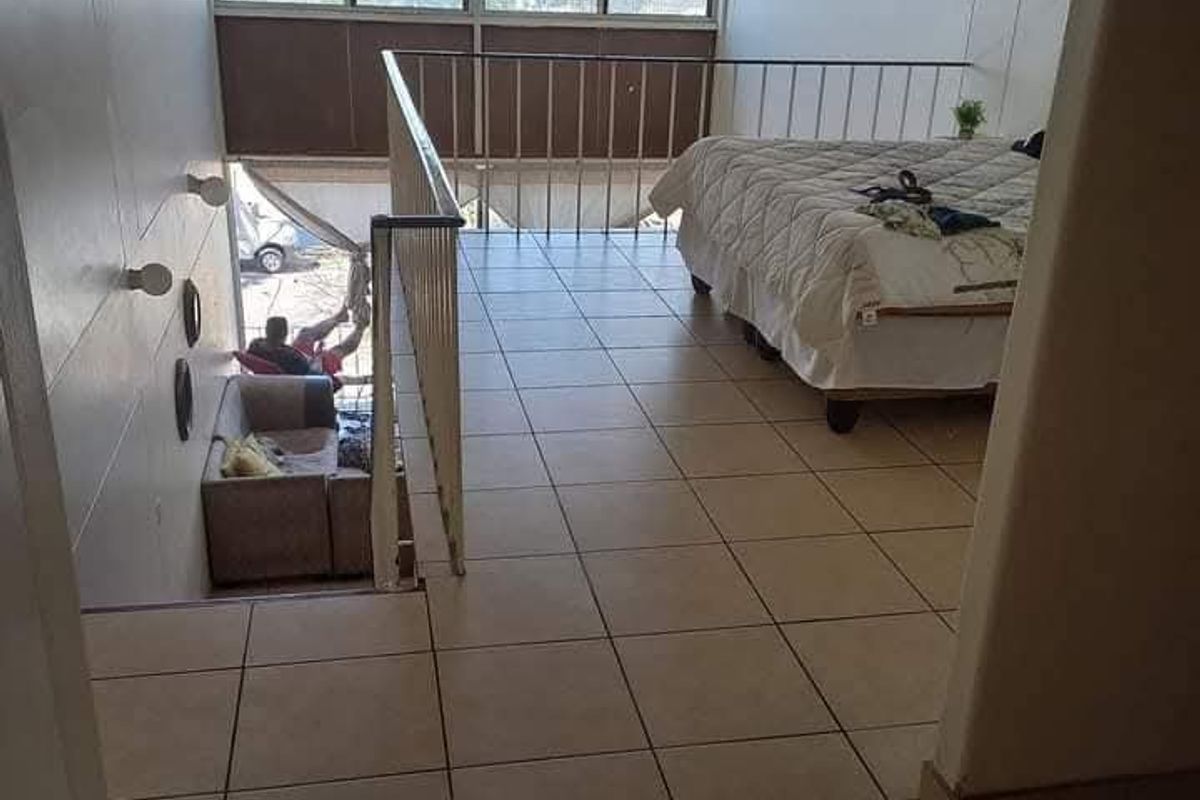 A mezzanine bedroom area featuring tiled flooring, a metal safety balustrade, and large windows overlooking the lower living space.