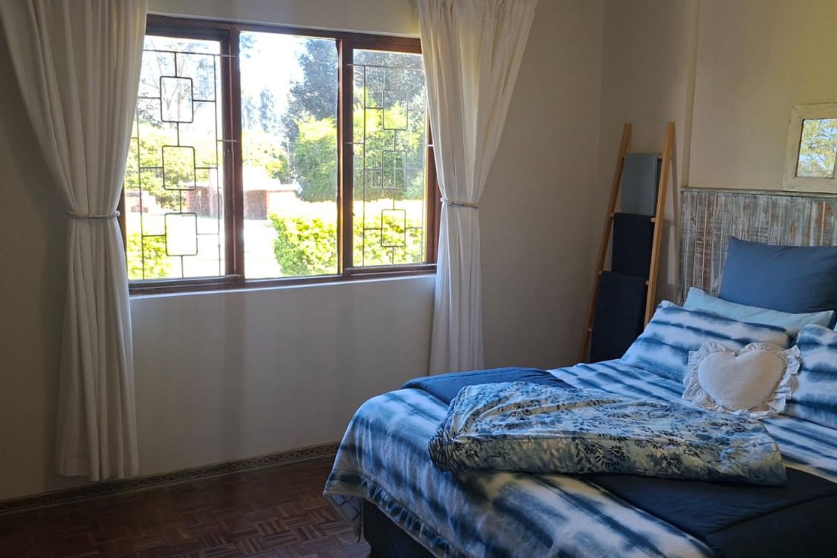 A cozy bedroom featuring parquet flooring and large windows with security bars, allowing for natural light and garden views.