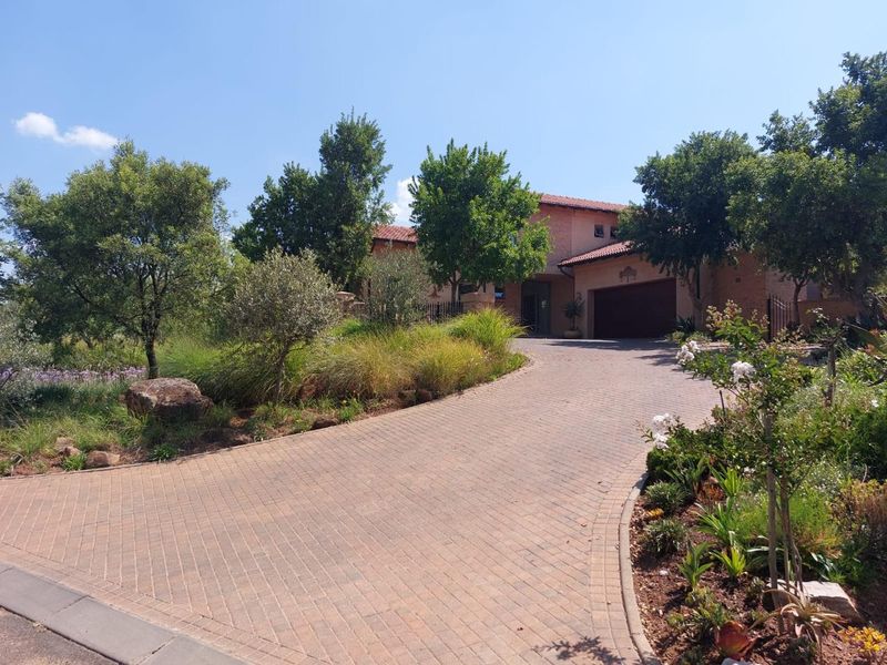 A well-maintained front elevation featuring a paved driveway leading to a double garage, surrounded by established landscaping and mature trees.