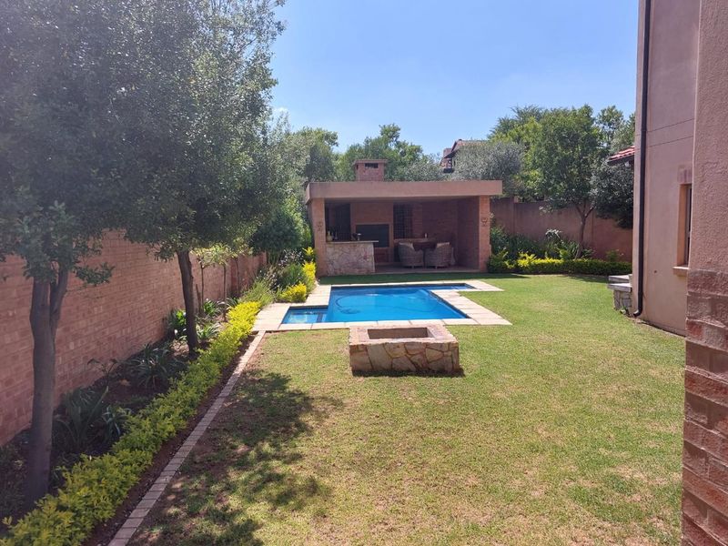 A well-maintained rear garden featuring a swimming pool, a stone-clad fire pit, and a covered patio area with a built-in braai.