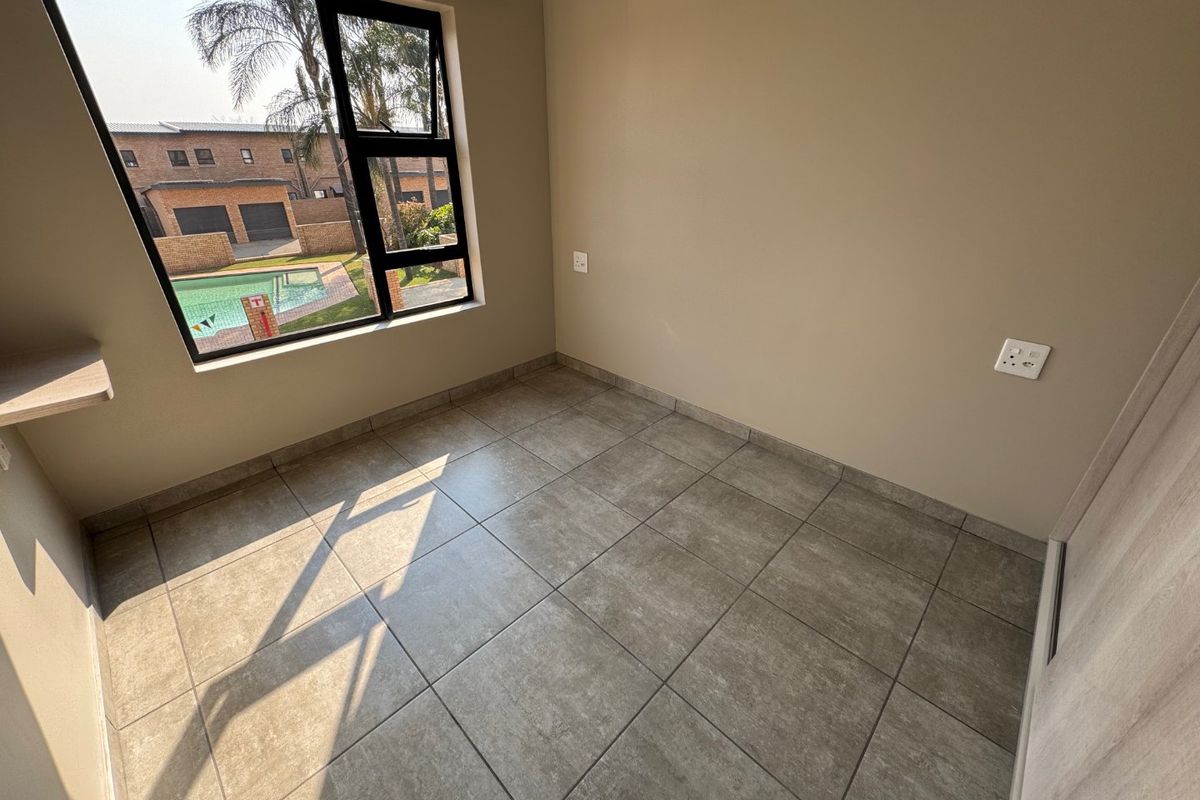 A bright, empty bedroom featuring neutral tiled flooring, a built-in cupboard, and a large window overlooking the complex pool area.