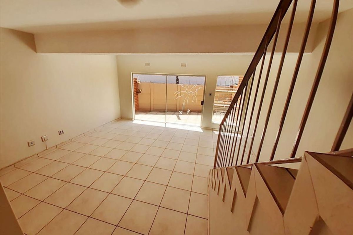 An empty, open-plan living area featuring tiled flooring, a staircase with a metal railing, and a sliding glass door leading to the exterior.