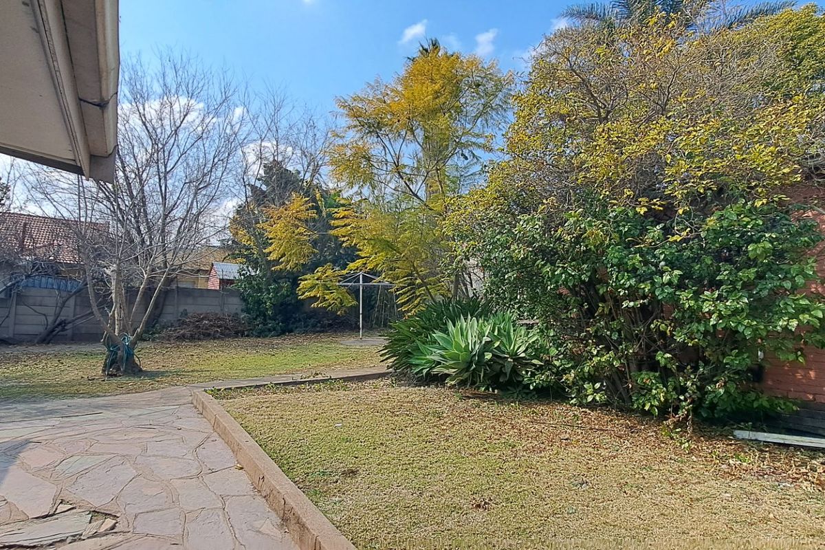 A spacious rear garden featuring a paved patio area, established trees, and a brick outbuilding, enclosed by a perimeter boundary wall.