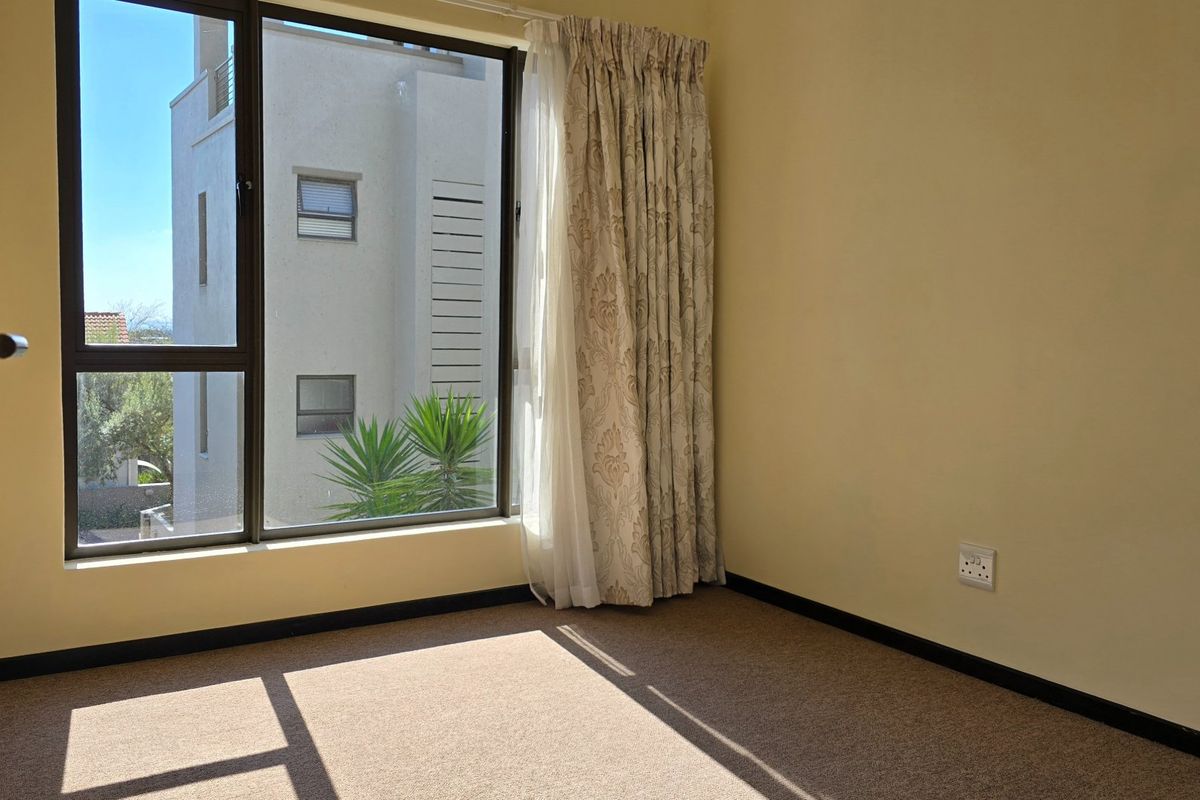 A bright, empty bedroom featuring neutral carpeting, dark skirting boards, and a large window providing natural light and an exterior view.