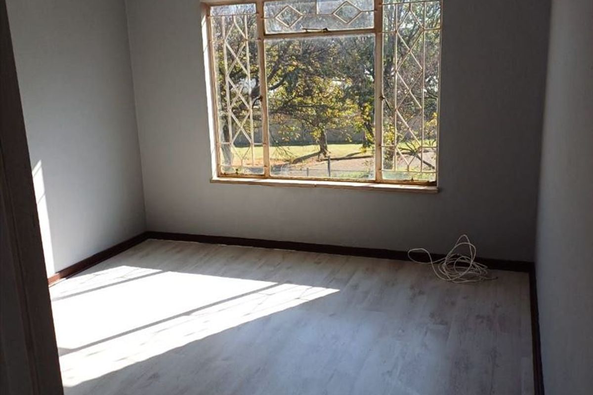 A bright, empty bedroom featuring light-toned laminate flooring, a ceiling fan, and a large window with security bars overlooking the garden.
