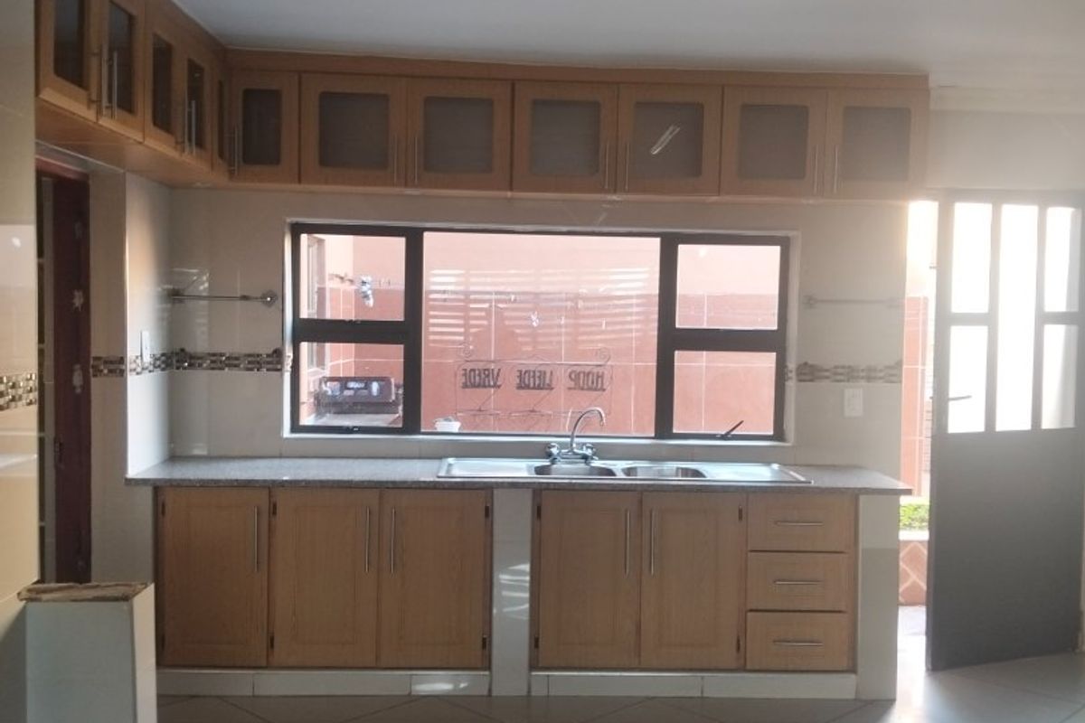 A clean, functional kitchen featuring built-in wooden cabinetry, a double basin sink, tiled backsplash, and neutral floor tiles with ample natural light.