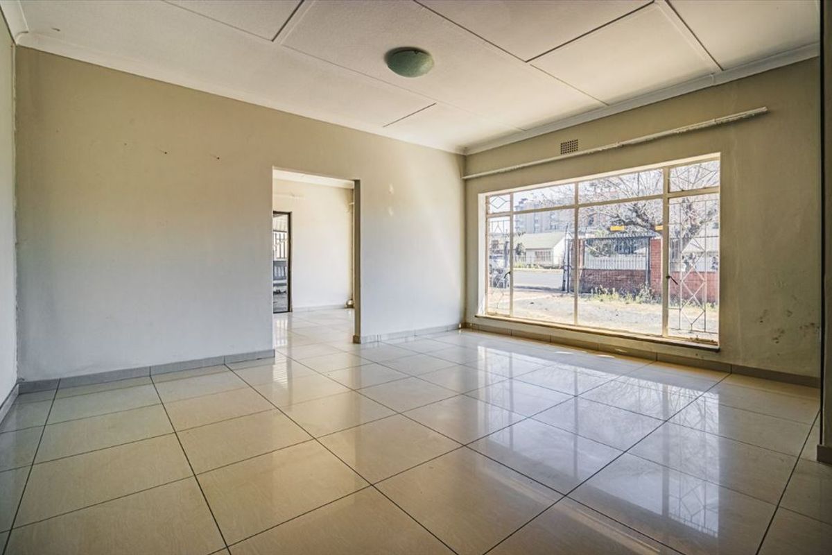 An empty, sunlit living room featuring large windows with security bars and neutral tiled flooring, offering a blank canvas for renovation.