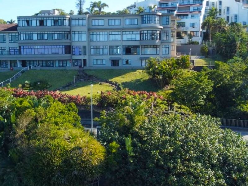 An elevated view of the apartment complex exterior, featuring a multi-story facade, landscaped grounds, and a swimming pool area.