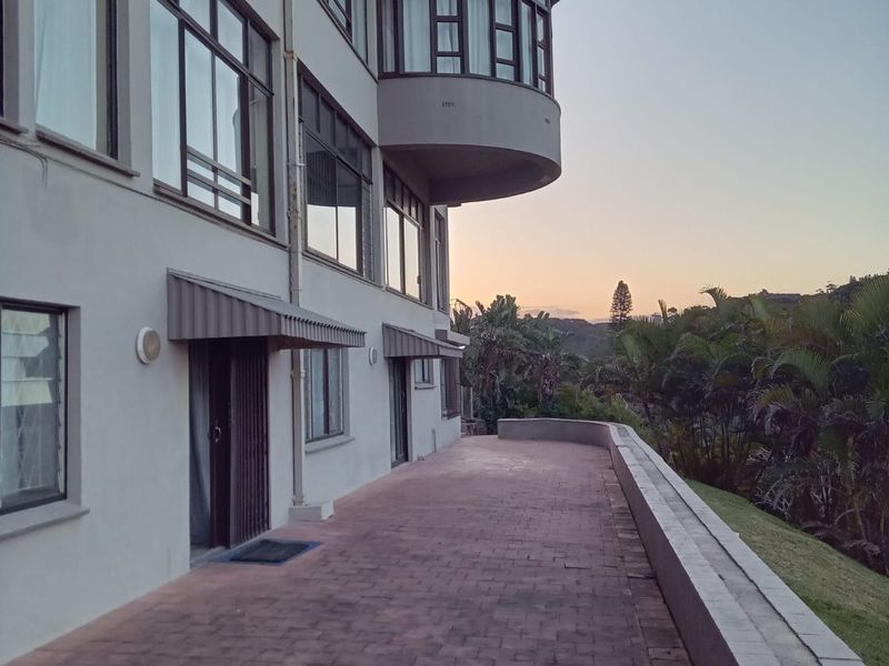 A spacious paved balcony terrace featuring a curved architectural design, awnings over doorways, and views of the surrounding landscape.