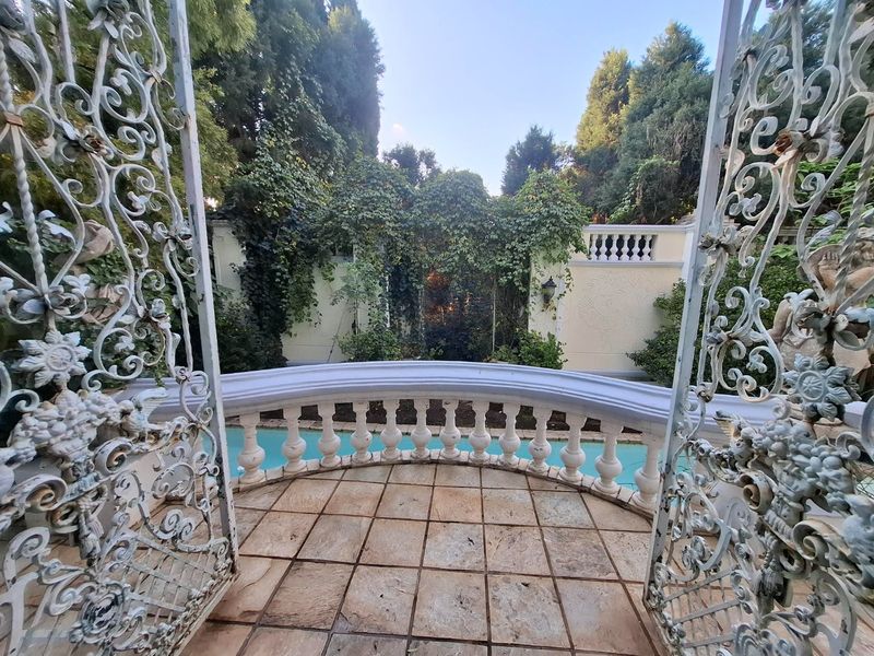 A charming outdoor pool area featuring a stone-tiled patio, decorative balustrade, and ornate wrought iron gates set against a lush, landscaped garden wall.