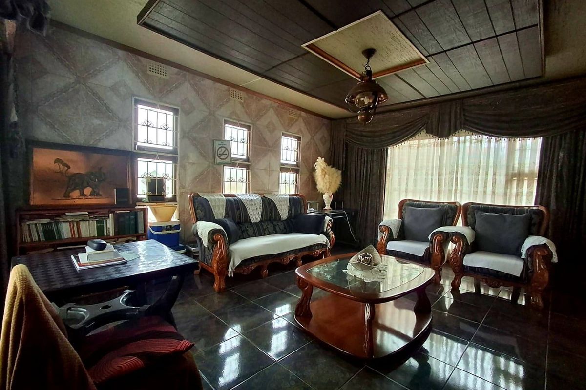 A spacious living room featuring dark tiled flooring, a coffered ceiling, and decorative wall finishes with large windows fitted with security bars.