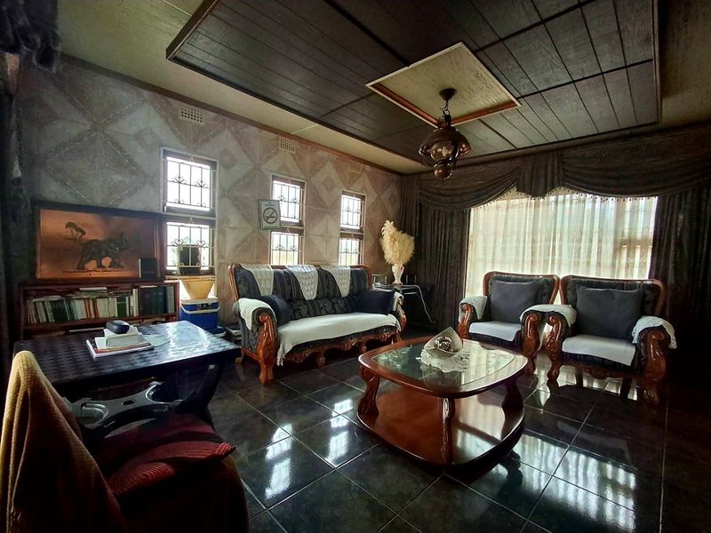 A spacious living room featuring dark tiled flooring, a coffered ceiling, and decorative wall finishes with large windows fitted with security bars.