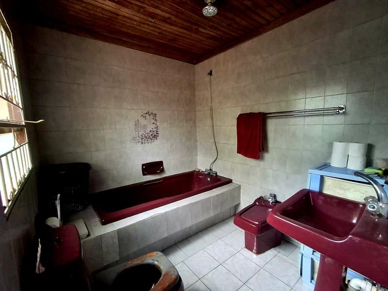 A dated bathroom featuring a burgundy suite, including a built-in bathtub, pedestal basin, bidet, and toilet, with tiled finishes and a timber ceiling.