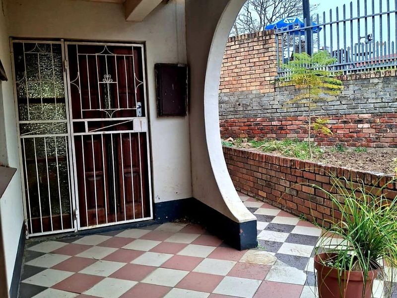 A covered patio area featuring checkered tile flooring, a decorative curved architectural pillar, and a secure entrance gate leading to a brick-walled garden space.
