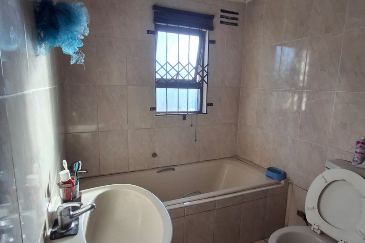 A dated bathroom featuring a pedestal basin, built-in bathtub, and toilet with full-height wall tiling and security bars on the window.