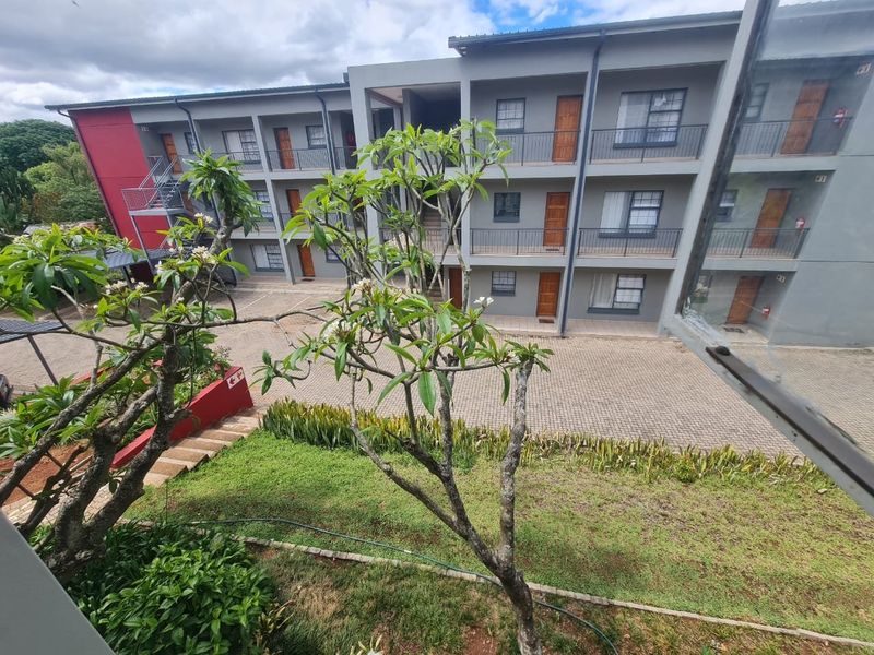 View of the apartment complex exterior, featuring a paved courtyard, landscaped garden area, and multi-level building facade with balcony access.