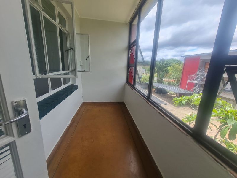 An enclosed balcony featuring concrete flooring and large metal-framed windows, offering an open view of the complex grounds.