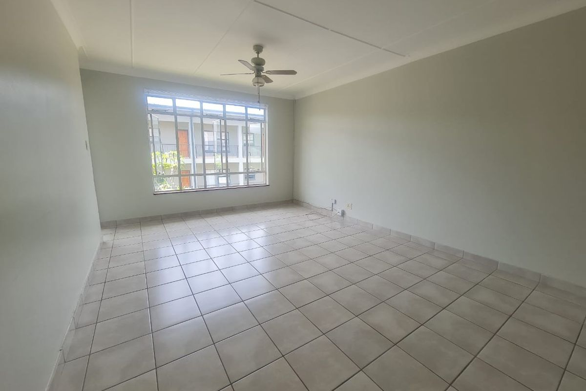 A spacious, empty living room featuring neutral tiled flooring, a ceiling fan, and a large window providing natural light.