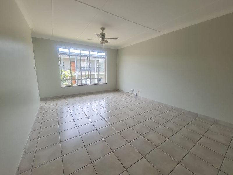 A spacious, empty living room featuring neutral tiled flooring, a ceiling fan, and a large window providing natural light.